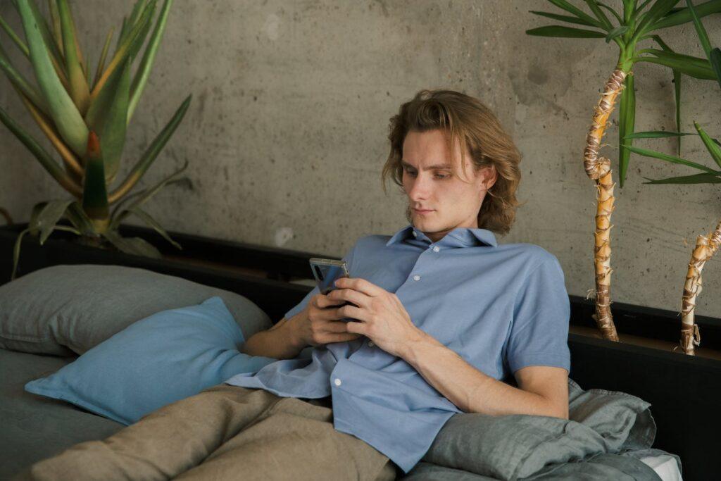 Young male in casual clothes lying on bed with smartphone and looking at screen