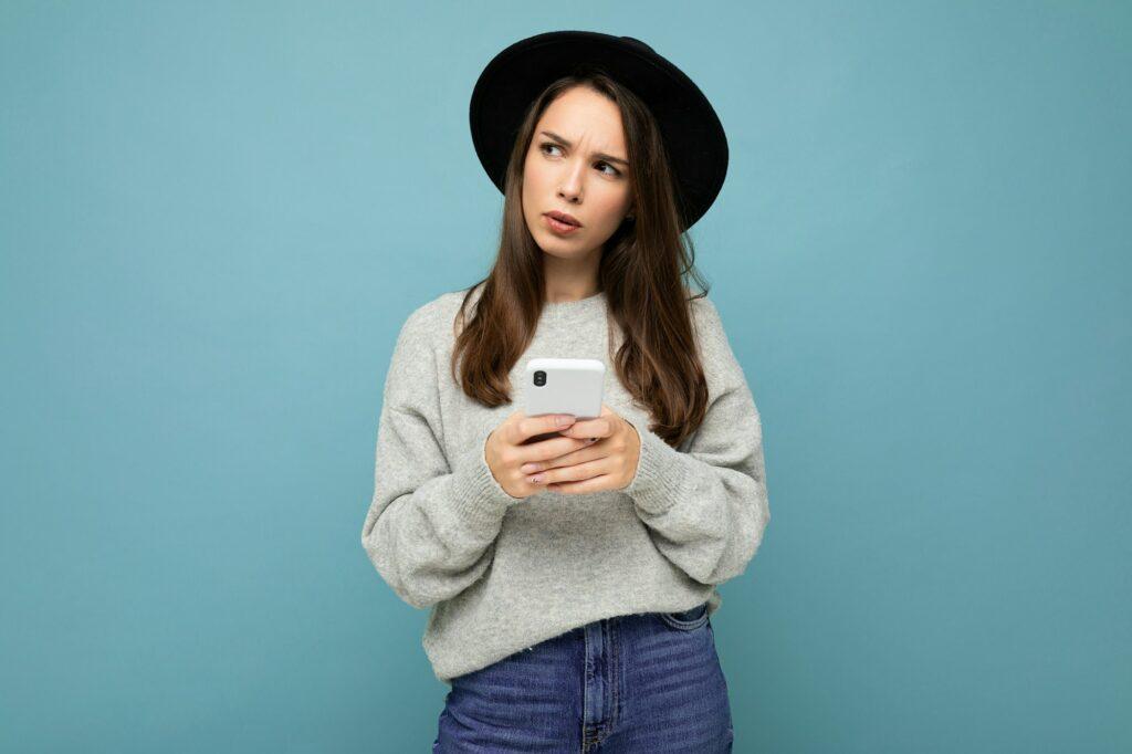 Beautiful young brunette woman thinking wearing black hat and grey sweater holding smartphone