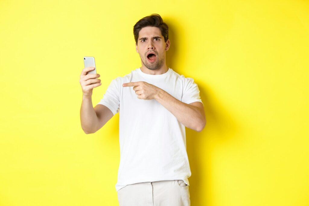 Confused man pointing finger at mobile phone screen, cant understand something, standing over yellow