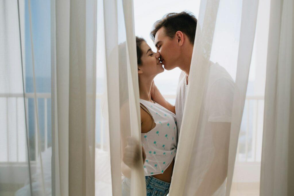 Graceful girl in white tank-top gently kissing her brunette boyfriend hiding behind light curtains.