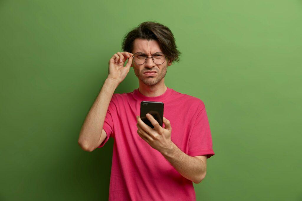 Serious man looks scrupulously through optical glasses, keeps hand on rim, uses mobile phone for onl